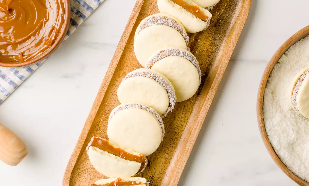 Alfajores redondos de masa suave color marfil rellenos de dulce de leche, bordes cubiertos con coco rallado dispuestos en una bandeja de madera