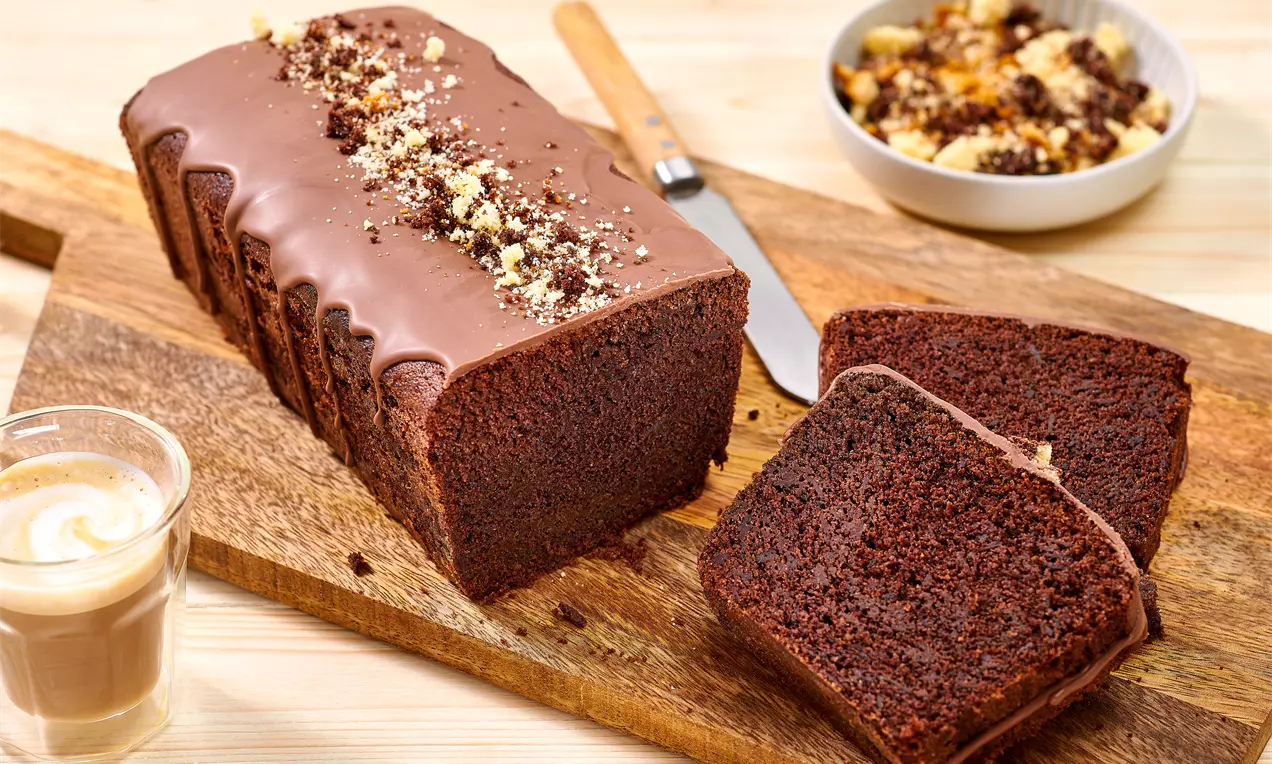 Chec dreptunghiular cu glazură lucioasă de ciocolată și firimituri crocante, feliat pe un platou de lemn lângă un pahar cu cafea cremă