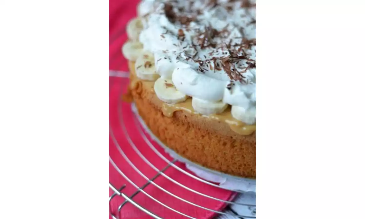 Banoffee cake met een goudbruine cakebodem, romige karamellaag, plakjes banaan, slagroomtoefjes en chocoladeschaafsel op een rooster