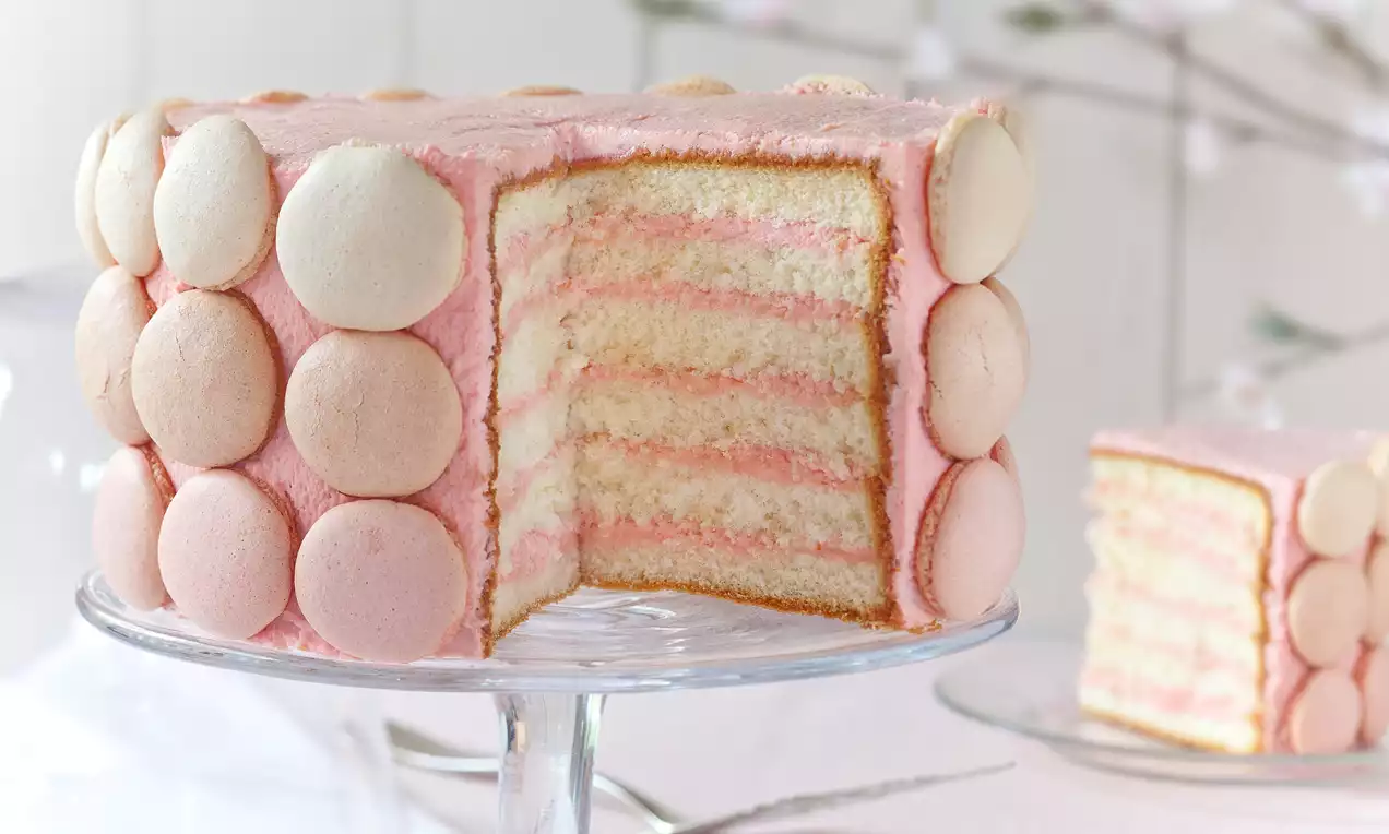 Wysoki tort warstwowy o miękkich różowo-biszkoptowych pasach, udekorowany kremem i pastelowymi makaronikami, na szklanej paterze
