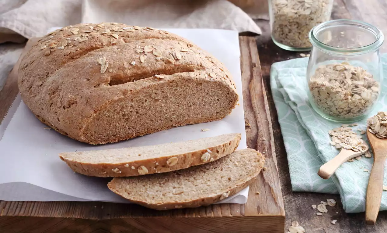 Gömbölyű zablisztes kenyér aranybarna héjjal, laza vágott szeletekkel, apró zabpehely szemekkel, rusztikus fatálcán fehér papírlapon