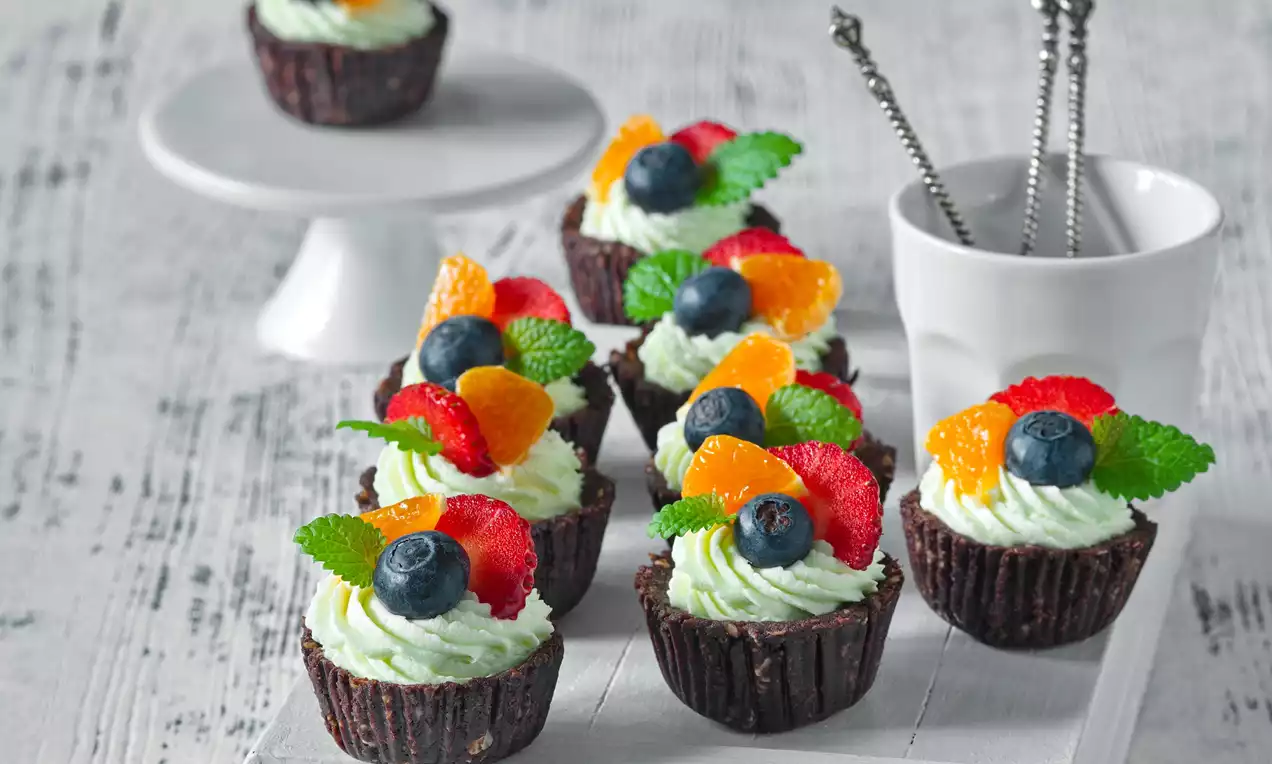 Čokoládové košíčky plněné světlým limetkovým krémem, zdobené borůvkami, plátky citrusů, jahodami a zelenými lístky na dřevěném podnose