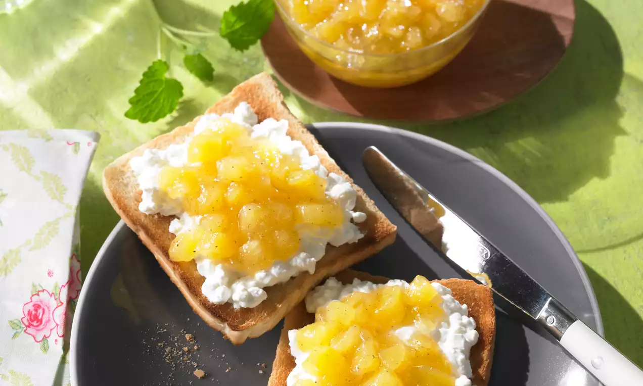 Goudgele ananasconfituur met glanzende stukjes op luchtige cottage cheese en geroosterd brood, gepresenteerd op een donkergrijs bord met een mes