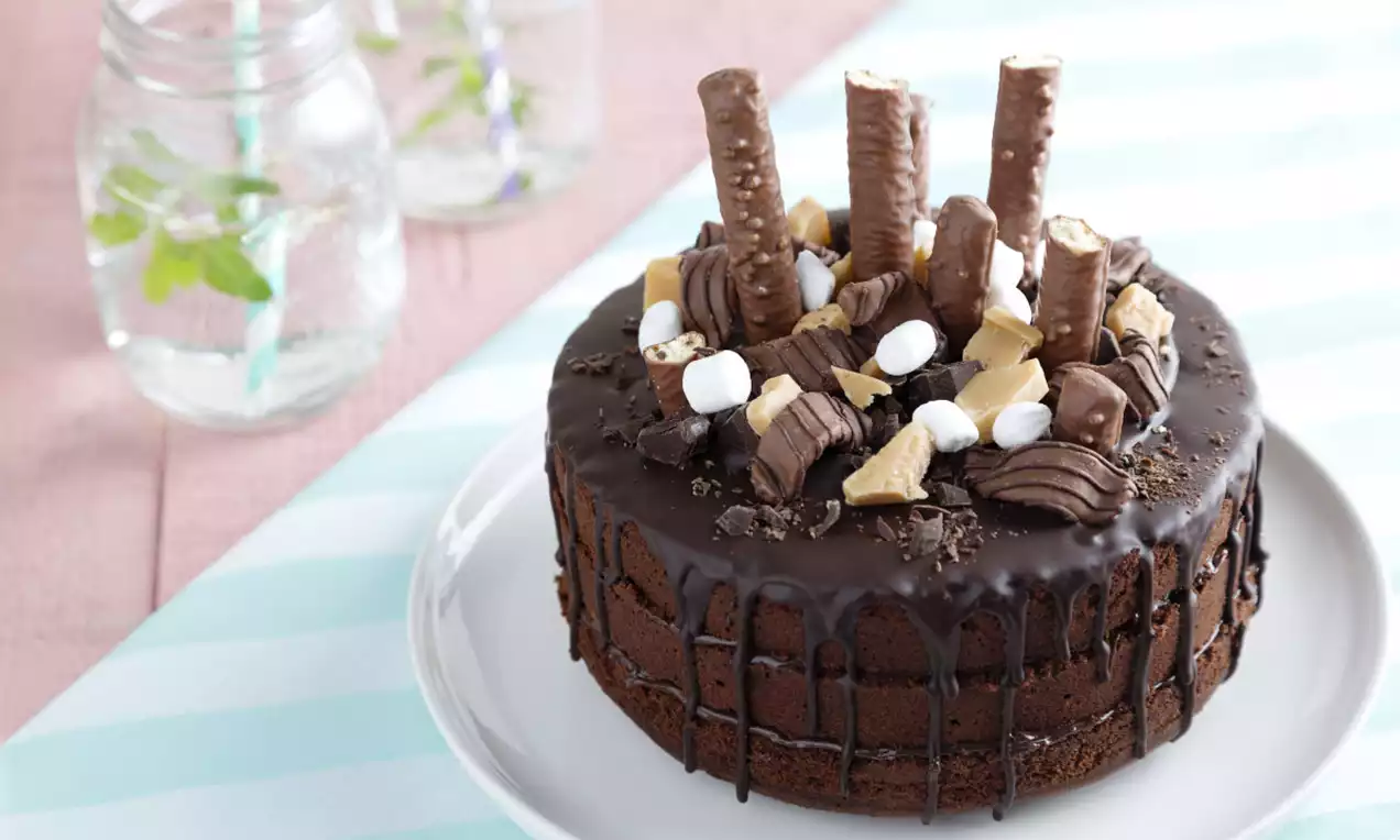 Ronde chocoladetaart met glanzende donkere ganache, gegarneerd met chocoladerepen, marshmallows en koekstukjes op een wit bord