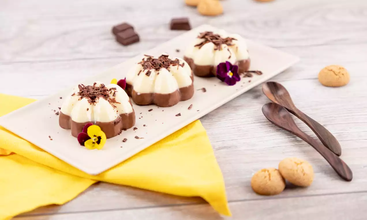 Pudding marmorat în straturi cremoase de ciocolată și vanilie, decorat cu fulgi de ciocolată și flori comestibile pe o farfurie albă rectangulară