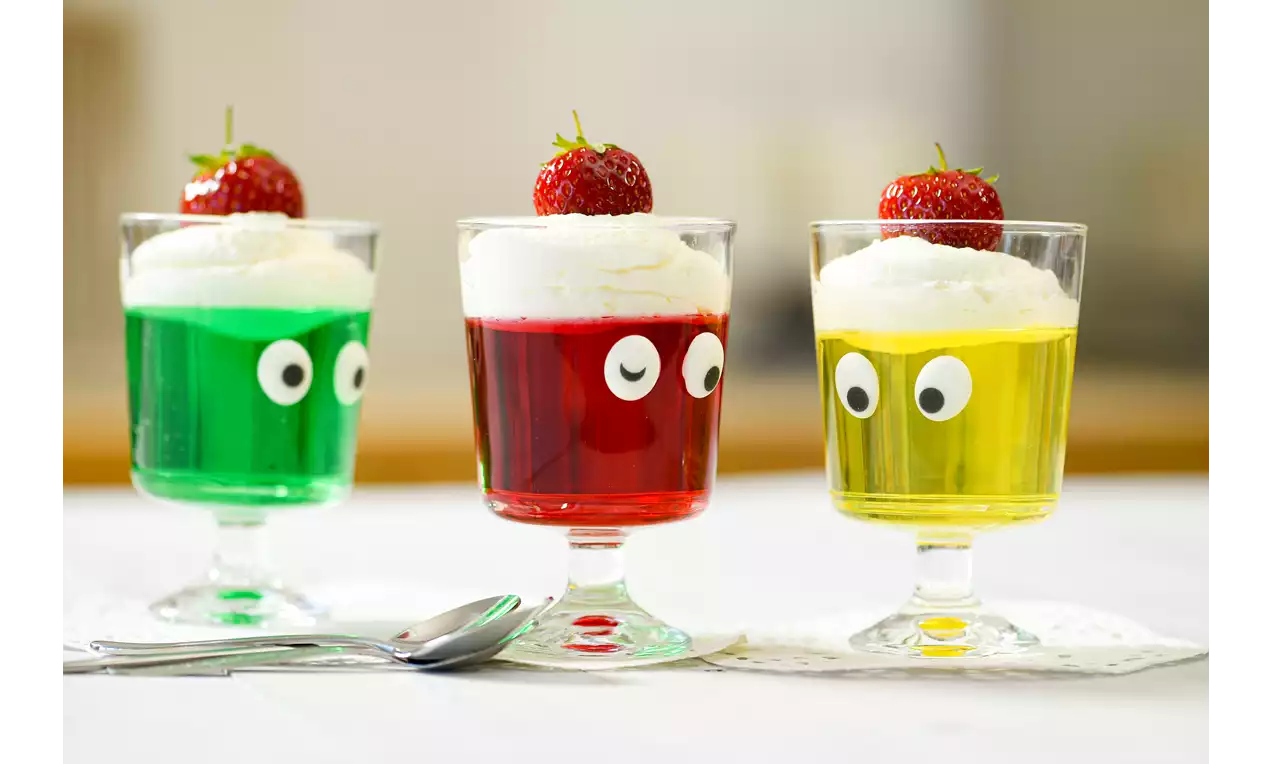 Drei Gläser mit Götterspeise in Grün, Rot und Gelb, Sahnetupf und Erdbeere obenauf, dekoriert mit Zuckeraugen auf hellem Tisch