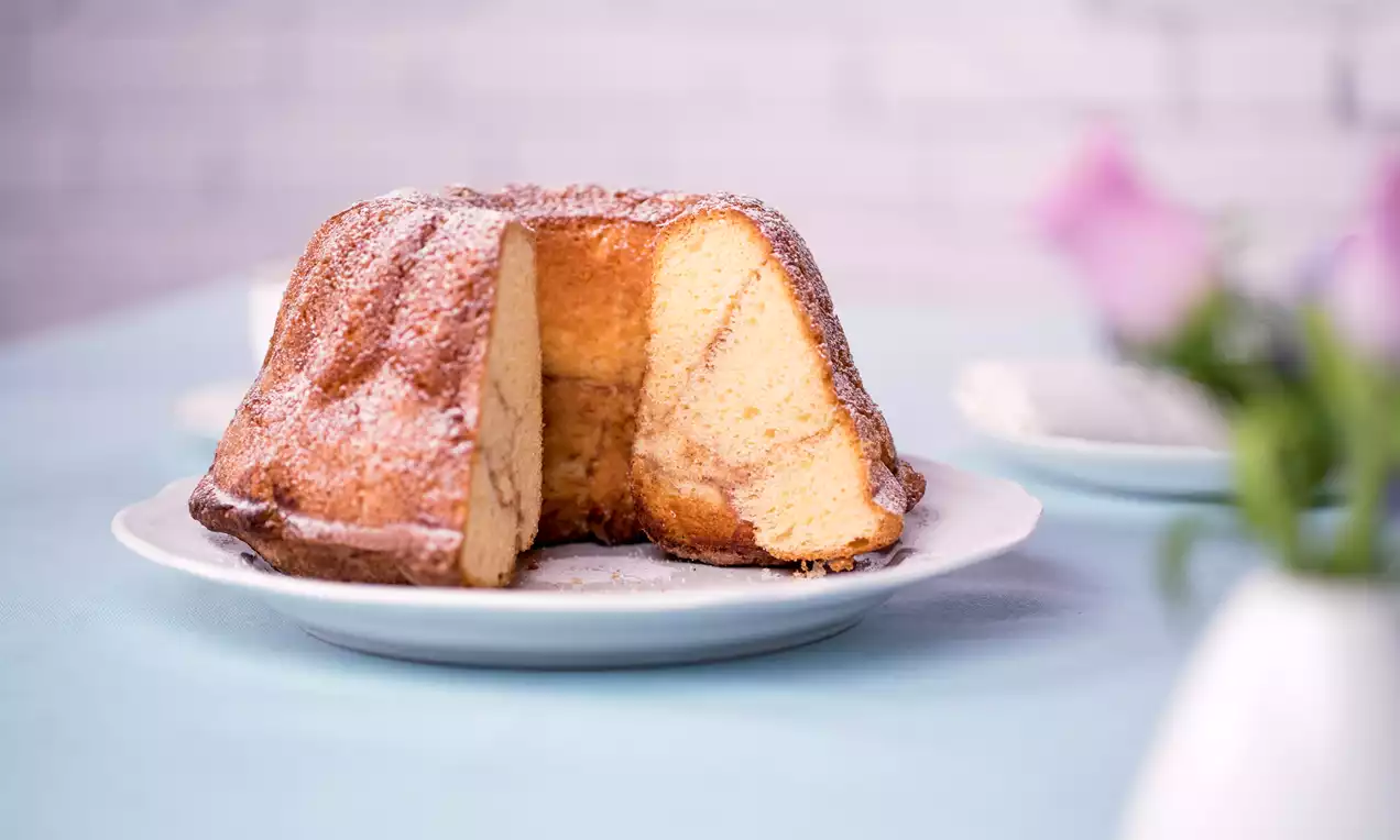 Goldbrauner Butter-Zimt-Gugelhupf mit sichtbarer Marmorierung und Puderzucker bestäubt auf weißem Teller, arrangiert auf hellblauer Tischdecke