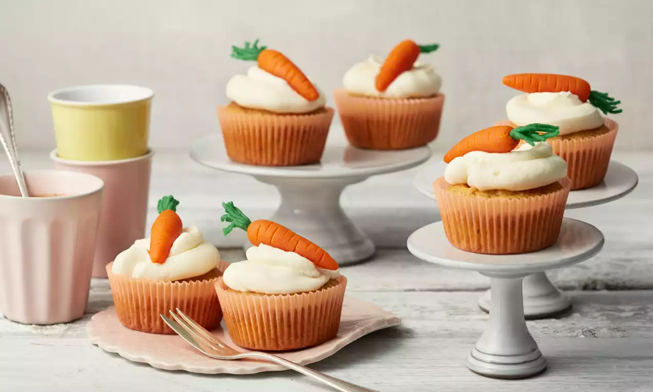 Zarte Rübli-Cupcakes in orangefarbenen Förmchen mit glatter Frischkäsehaube und dekorativen Marzipanmöhren auf kleinen weißen Tortenständern