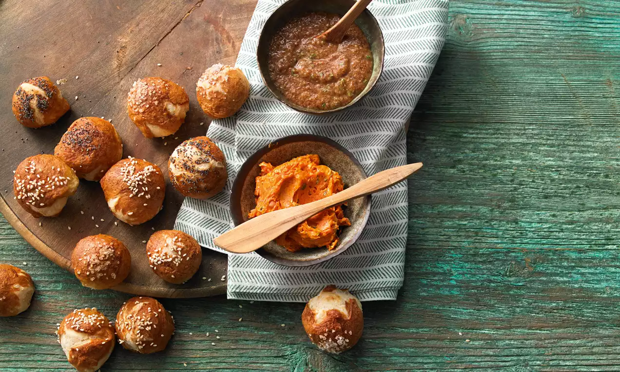 Kleine, goldbraune Bretzel-Bites mit Salz bestreut, neben Schalen mit cremigem Dip und leuchtender Tomatenbutter auf rustikalem Holzteller
