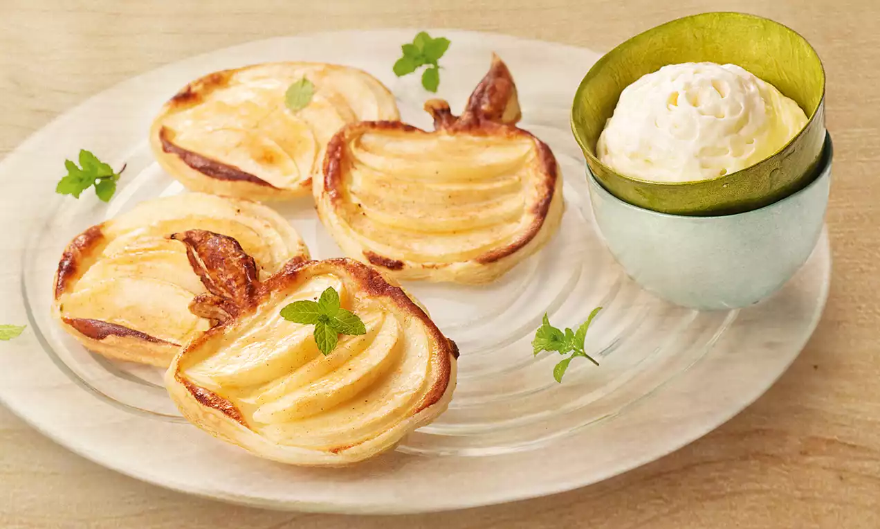 Petits feuilletés dorés en forme de pommes garnis de tranches d’apples brillantes et de feuilles de menthe sur une assiette transparente