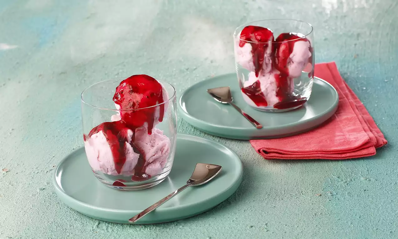 Cam bardakta pembe çilekli dondurma, parlak kırmızı sos ile süslenmiş, yeşil tabakta küçük kaşık ve pembe kumaşla sunulmuş