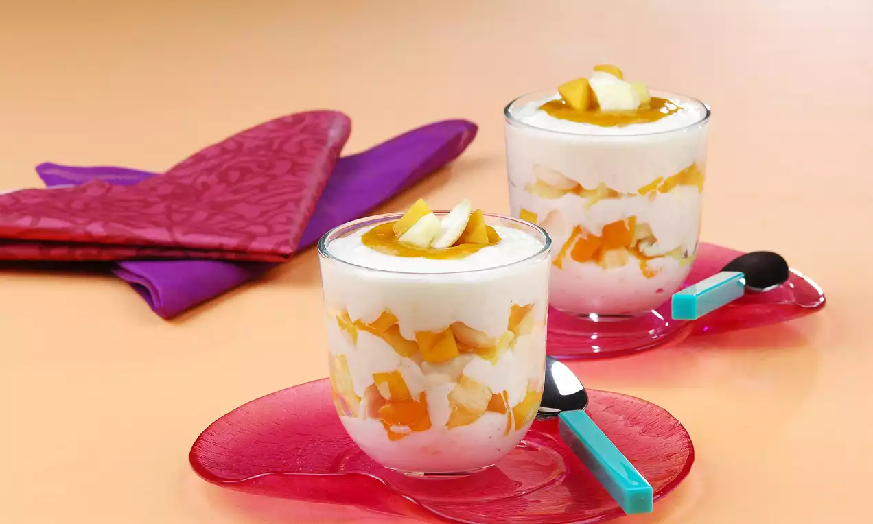 Şeffaf cam bardaklarda beyaz sütlaç, tropik meyve küpleri ve altın rengi sosla katmanlanmış, pembe tabakta mavi detaylı kaşıklarla sunulmuş