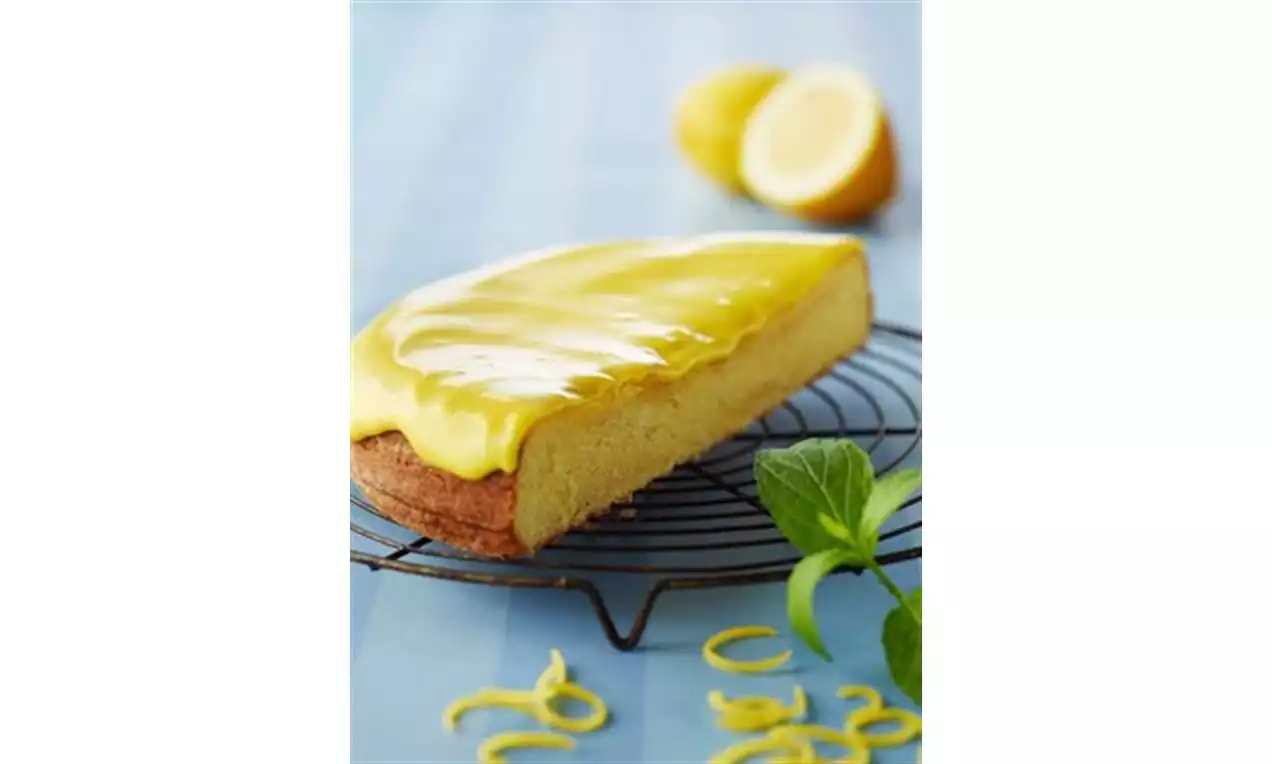 Avlang kake med gyllen sitronglasur på rist, lysegul inni med brun kant, omgitt av sitronskallstrimler, mynteblader og halve sitroner