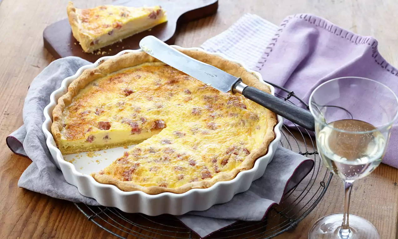 Goldgelbe Quiche Lorraine mit knusprigem Rand in einer weißen Form, auf einem Holzbrett mit einem Stück und einem Glas Weißwein daneben