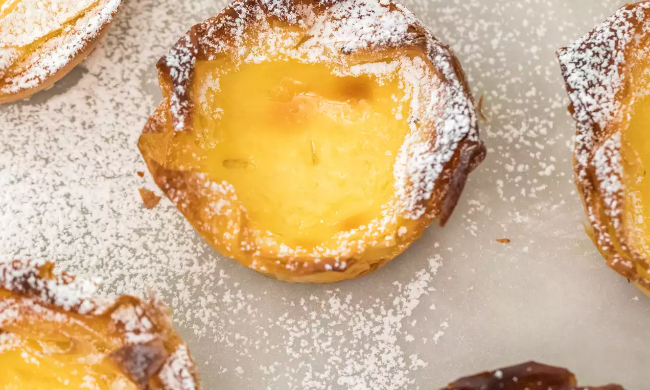 Pasteis de Nata con relleno dorado y brillante, borde crujiente y quemado, espolvoreados con azúcar glas sobre papel blanco