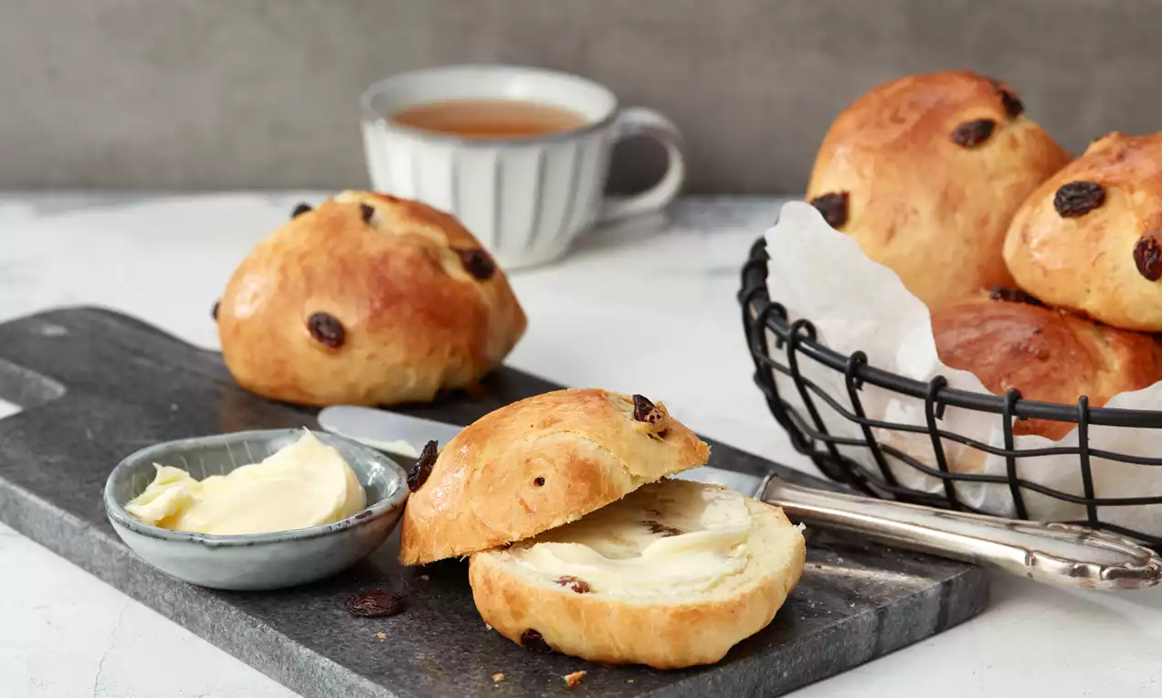 Ein goldbraunes Rosinenbrötchen mit weicher, glatter Textur, aufgeschnitten und mit Butter bestrichen, neben einem Messer auf dunklem Brett