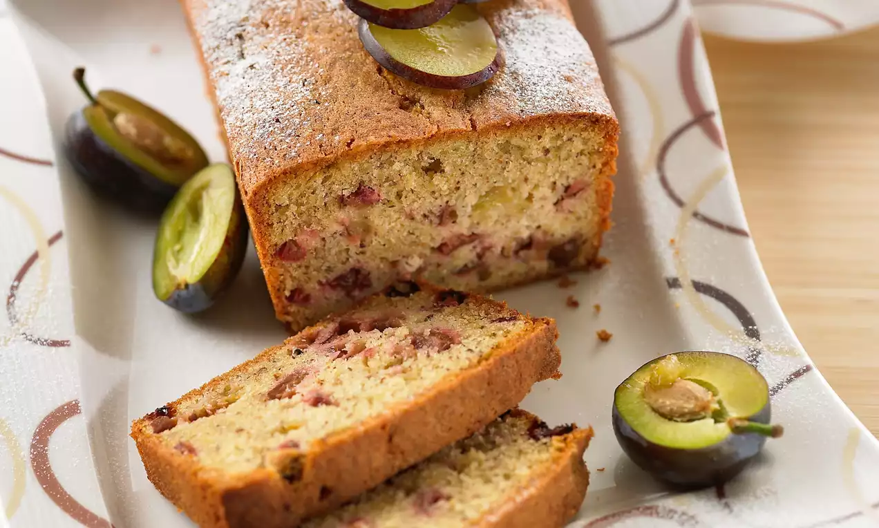 Cake doré aux prunes violettes, tranches dévoilant une texture moelleuse marbrée de morceaux fruités, présenté sur un plat clair avec des prunes fraîches