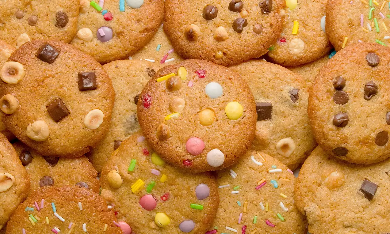 Galletas doradas redondas con bordes ligeramente crujientes, decoradas con chispas de chocolate, frutos secos, confites y sprinkles de colores