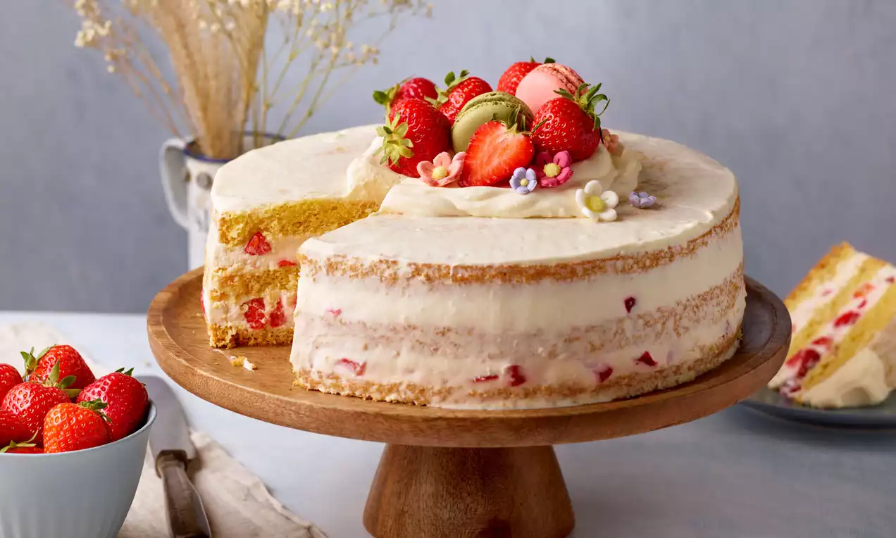 Naked Cake mit hellen Böden, Schichten aus Creme und Erdbeerstücken, garniert mit Erdbeeren, Macarons und Blüten auf einer Holzplatte