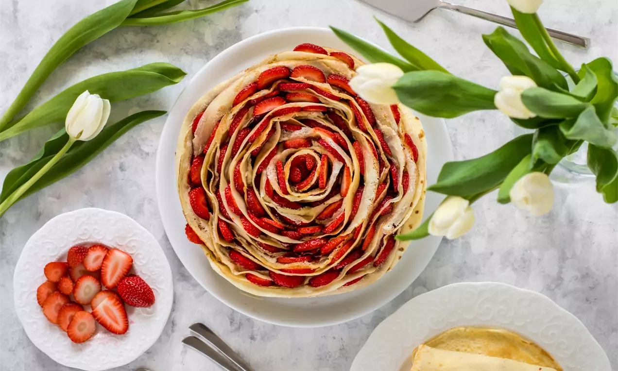 Торта от тънки, светложълти палачинки, подредени в кръгли пластове като разцъфнало цвете, гарнирана с нарязани на филийки яркочервени ягоди