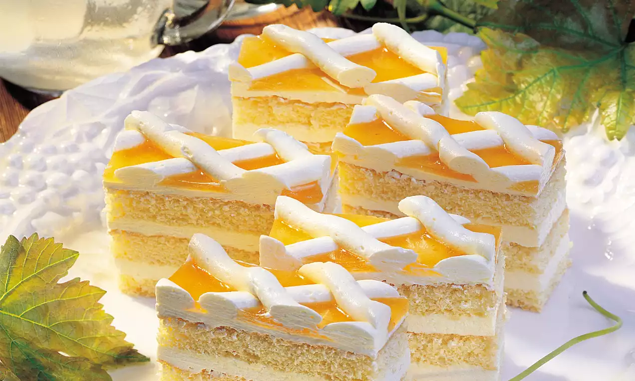 Wachauer Schnitten mit hellgelbem Biskuit, Schichten aus heller Creme, goldgelber Glasur und weißer Zuckergitterdekoration auf weißer Platte