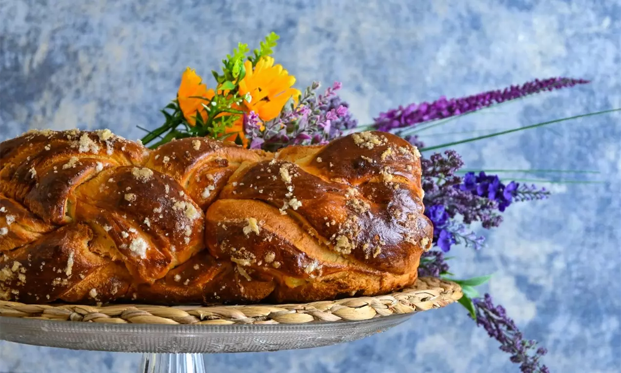 Златисто-кафяв, плитен козунак с разтегливи конци и нежен филинг, поръсен с едри захарни кристали, върху плетена подложка и свежи цветя
