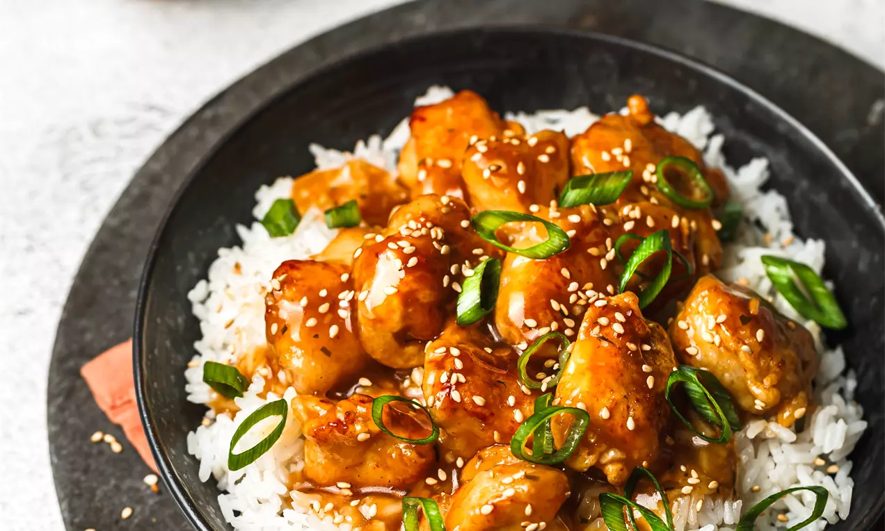 Picture - Glossy orange-glazed chicken pieces topped with sesame seeds and sliced green onions on fluffy white rice in a black bowl