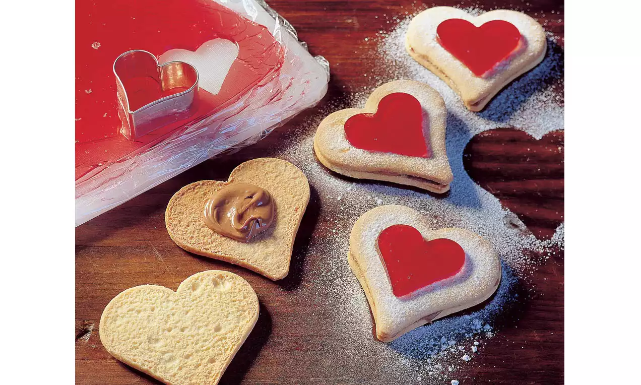 Brosse hartvormige koekjes met een glanzend rood laagje, poedersuiker en een dot chocolade op een houten ondergrond met uitsteekvorm