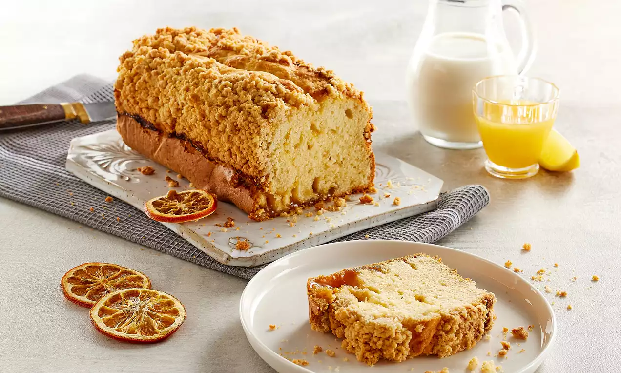 Altın sarısı crumble kaplı, içi yumuşak limon soslu kek, beyaz tabakta bir dilimle kuru limon dilimleri ve içecekler eşliğinde sunulmuş