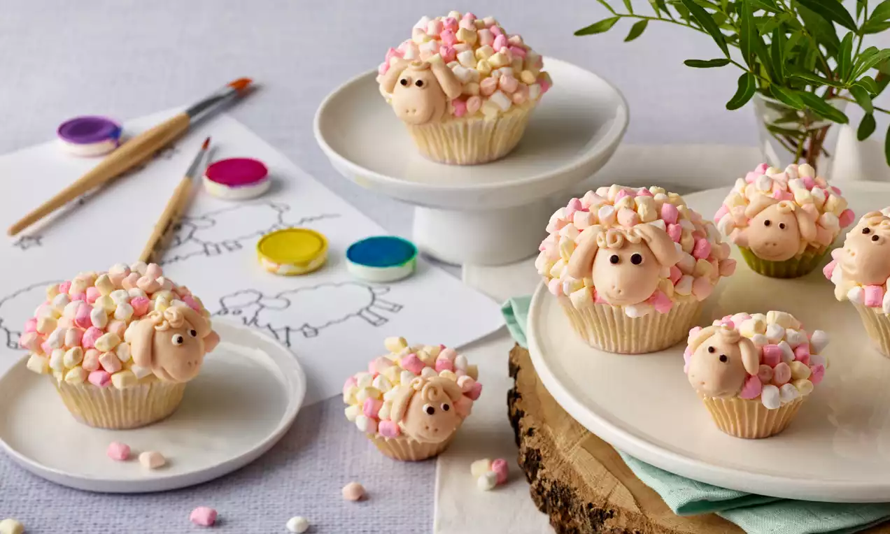 Vanillefarbene Cupcakes dekoriert mit bunten Mini-Marshmallows und Zuckerköpfchen in Schafform auf Tellern und Holzbrett arrangiert
