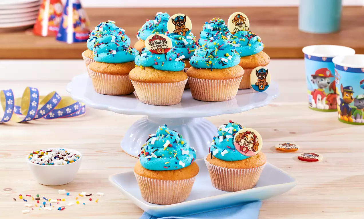 Cupcakes mit goldenem Teig und wolkenförmiger blauer Creme, bestreut mit bunten Streuseln und PAW Patrol-Dekoren auf weißem Geschirr