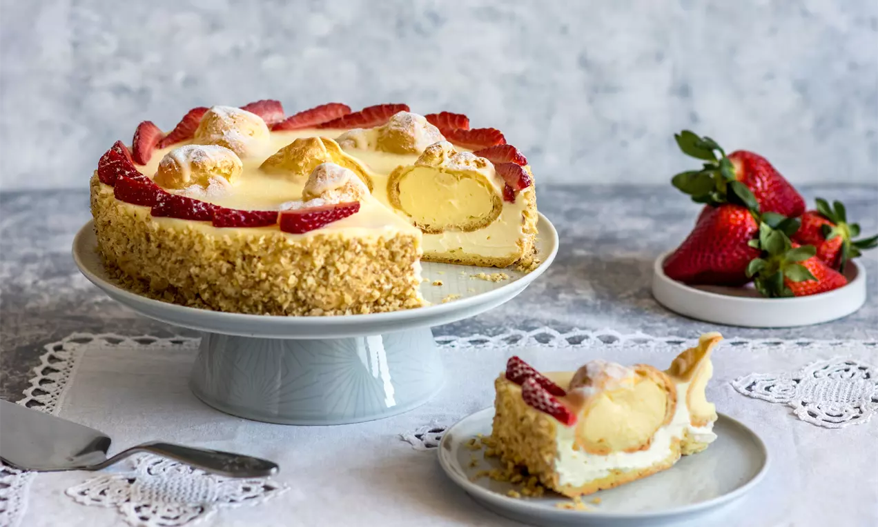 Кръгла еклерова торта с кремообразен ванилов мус, украсена с еклери и свежи резени ягоди, поставена върху бяла порцеланова стойка