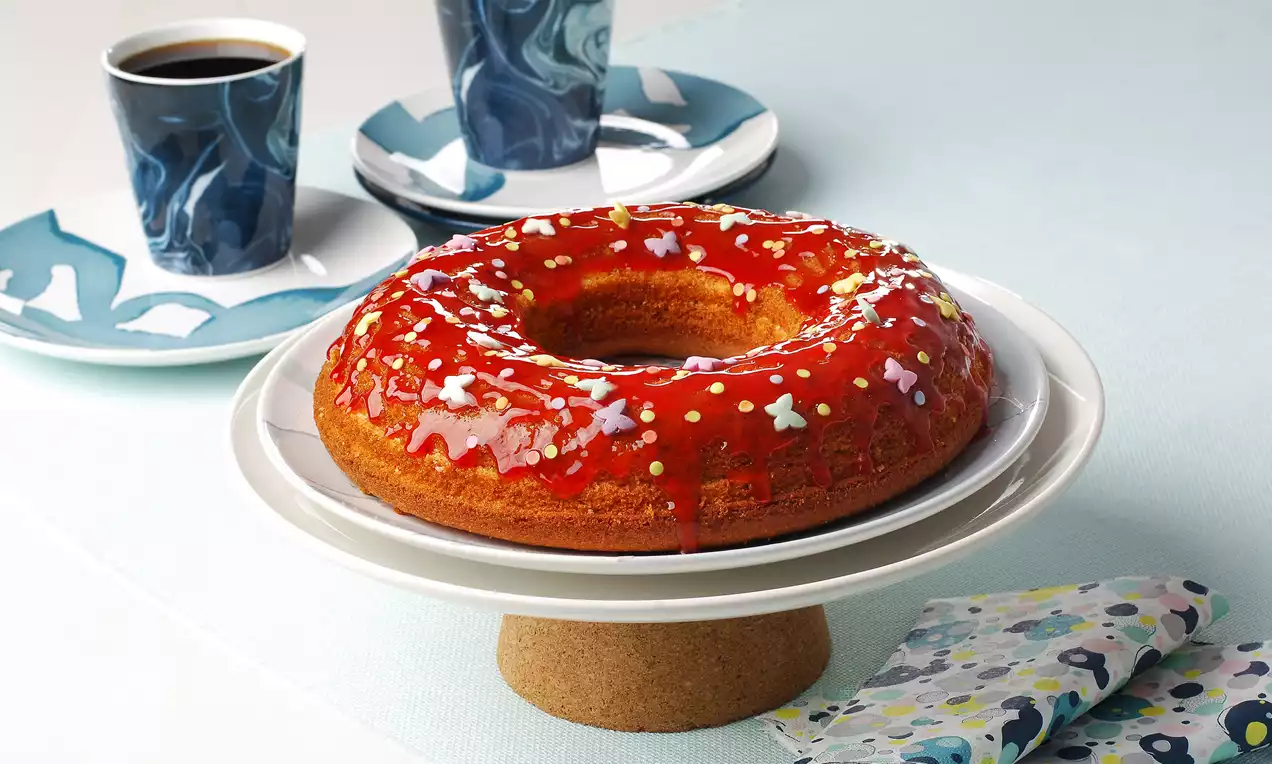 Pembe parlak glazür kaplı halka kek, renkli şekerlemelerle süslenmiş, beyaz ayaklı tabakta, yanında desenli fincan ve peçete