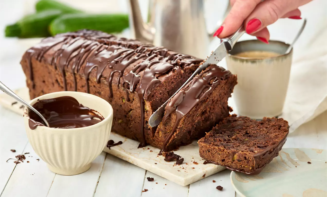 Saftiger Zucchini-Schoko-Kuchen in Kastenform mit glänzender Schokoglasur, angeschnitten auf Holzbrett neben einer Schüssel mit flüssiger Schokolade