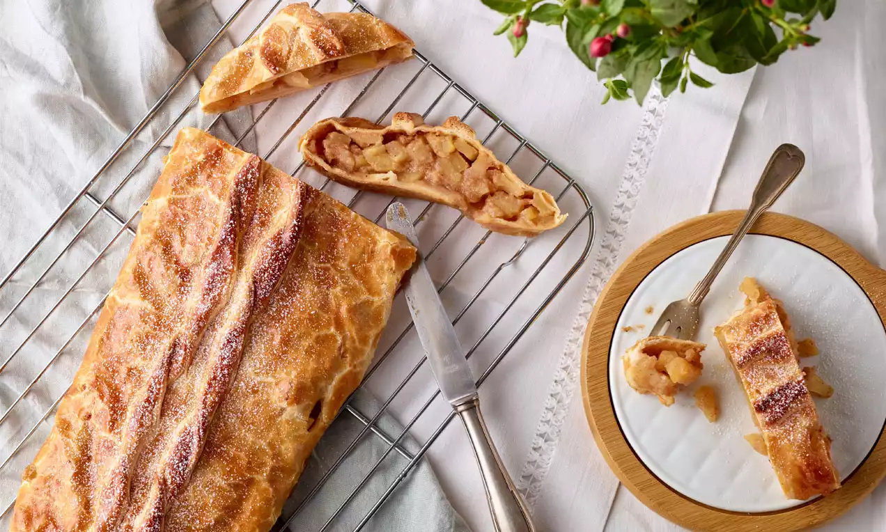 Goldbrauner, glatter Apfelstrudel aus Blätterteig mit feinen Zuckerkristallen, angeschnitten auf einem Gitter neben Teller und Besteck