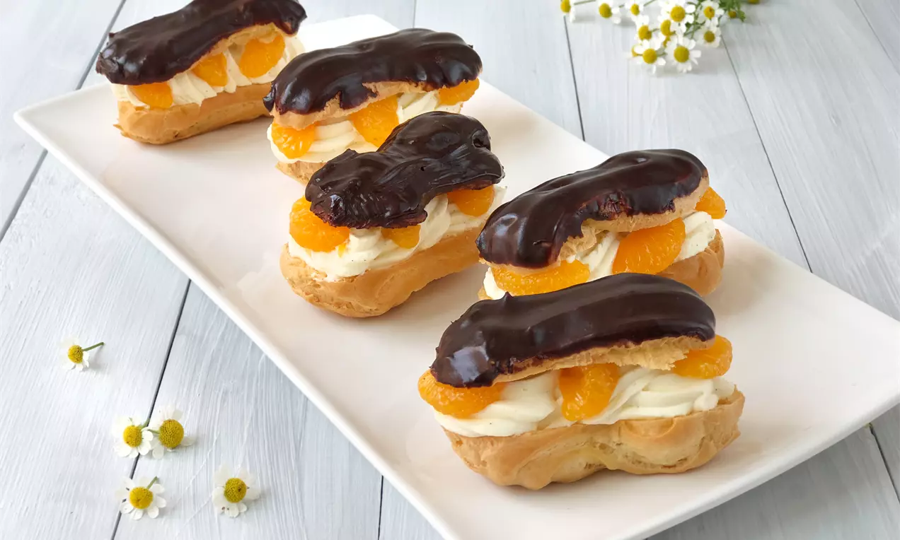 Eclairs mit goldbraunem Brandteig, gefüllt mit Vanille-Sahne und Mandarinen, überzogen mit glänzendem dunklem Schokoladenüberzug auf weißer Platte
