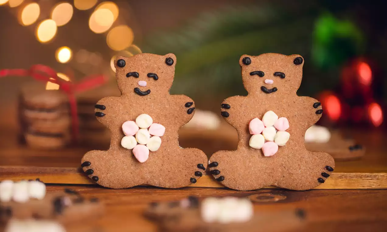 Dwa brązowe pierniczki w kształcie misiów z czarnymi detalami i brzuszkami z miniaturowych różowo-białych pianek na drewnianym tle z rozmytymi świątecznymi ozdobami w tle