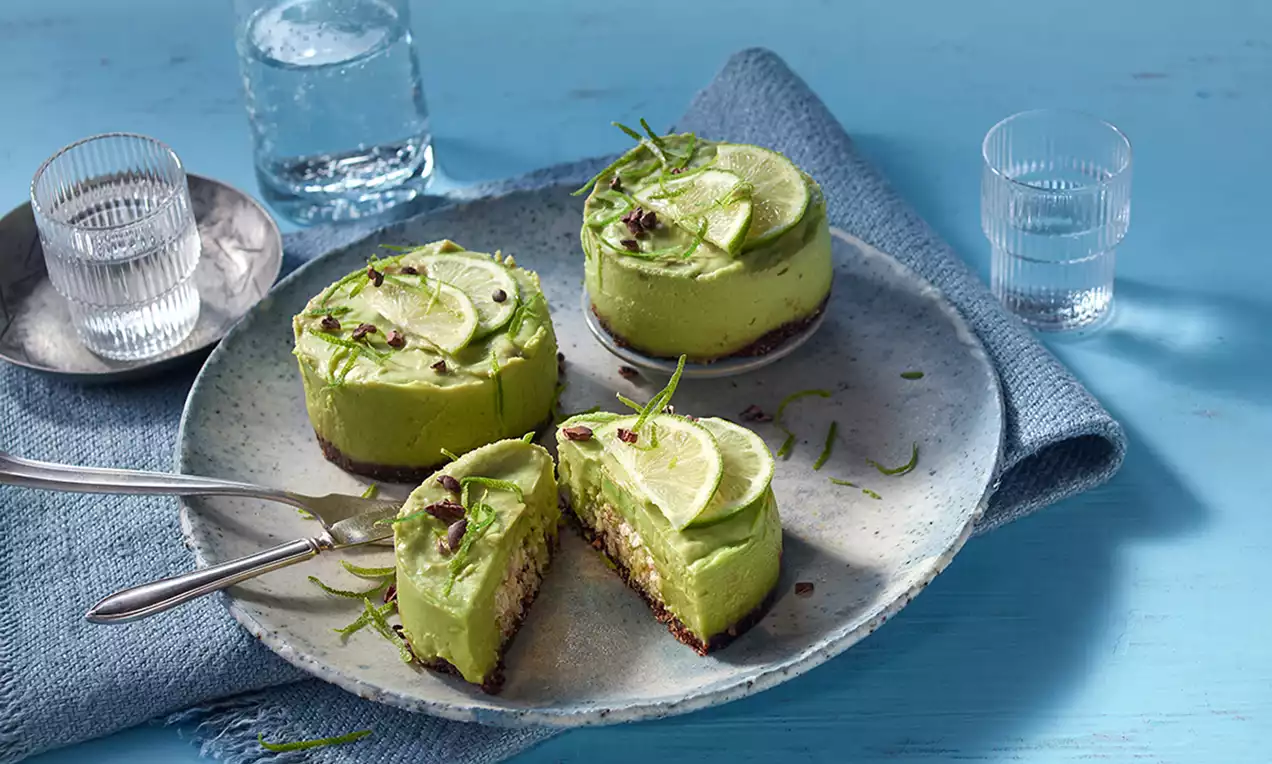 Mini prăjituri raw cu cremă verde de avocado și lime pe blat crocant, decorate cu felii de lime, coajă rasă și fulgi de cacao pe o farfurie în nuanțe de bleu