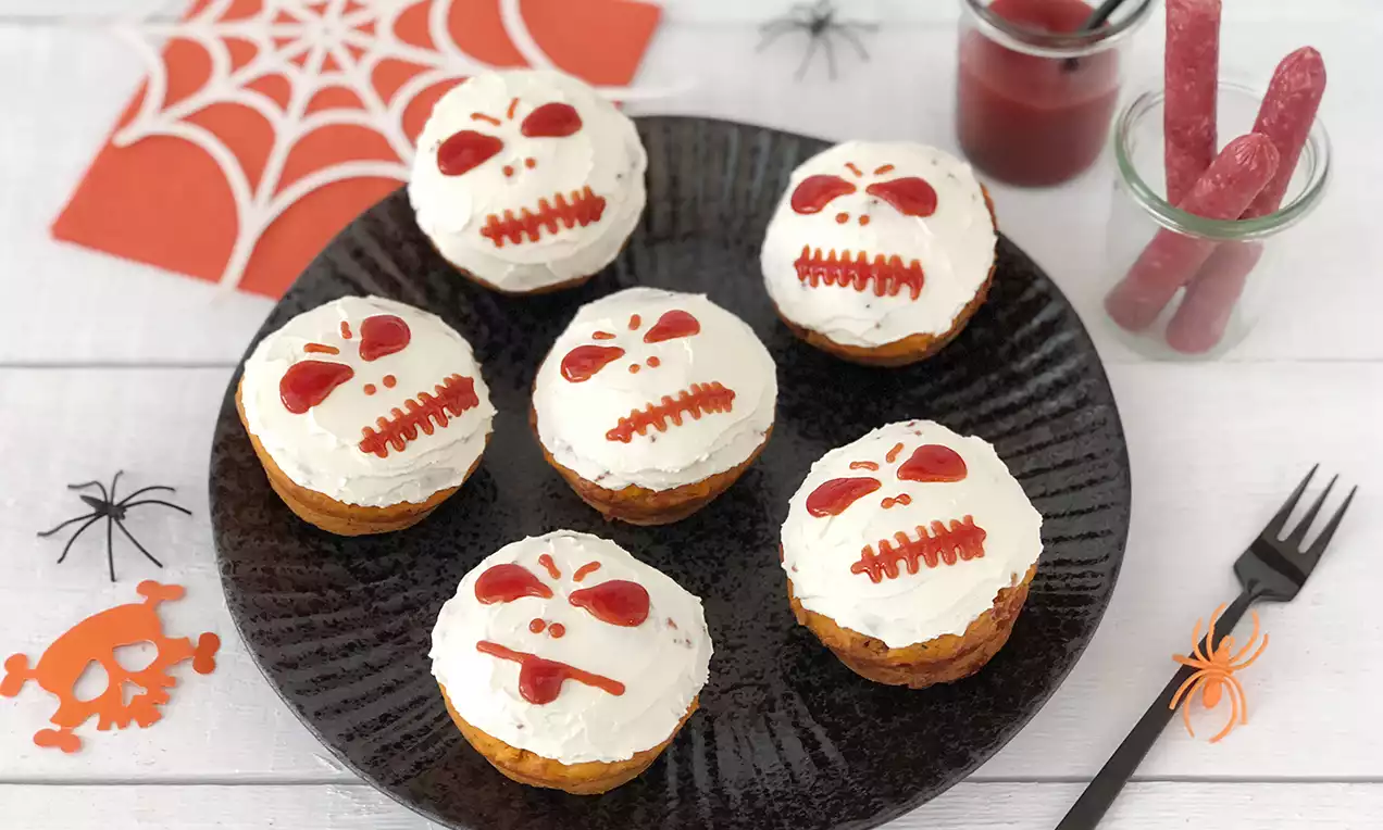 Sechs Muffins mit weißer Creme und roten Skelett-Gesichtern, angerichtet auf schwarzem Teller, umgeben von Halloween-Dekorationen