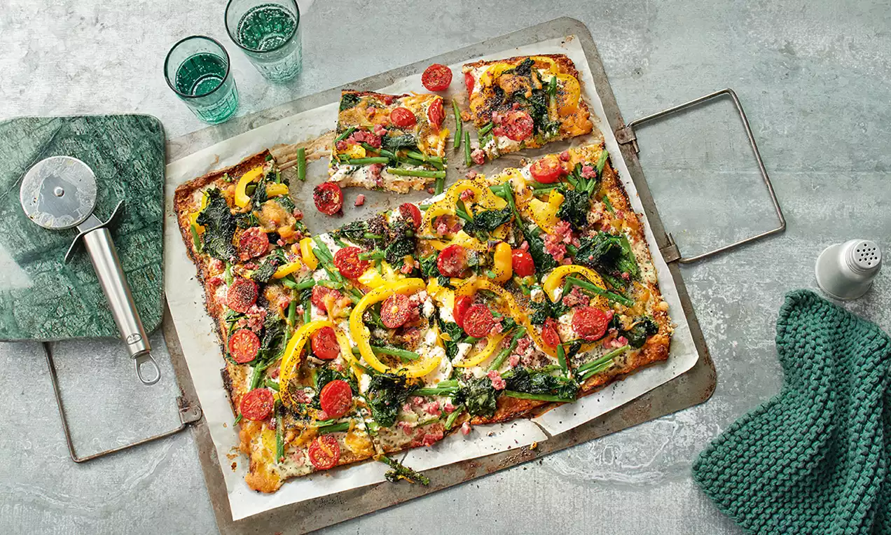 Bunte Pizza mit Blumenkohlboden, saftigem Gemüse wie gelber Paprika, grünen Brokkoliröschen und roten Tomaten auf Backpapier serviert