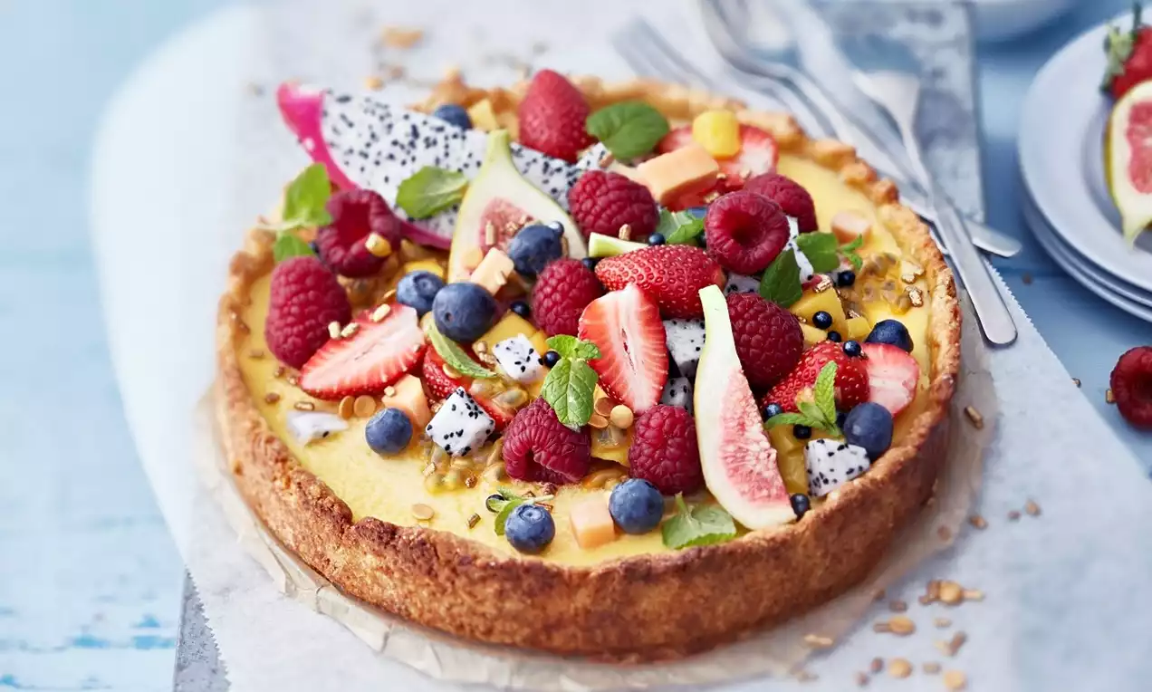 Kesäinen sitruunapiirakka kulmikkaalla keksipohjalla, täytteen päällä värikäs asettelu tuoreita marjoja, hedelmiä ja mintunlehtiä