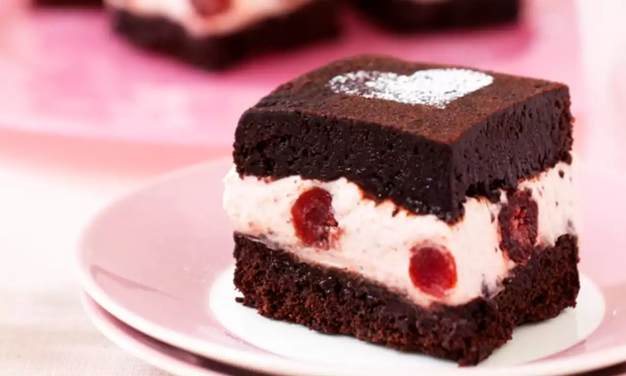 Bruin vierkant taartje van zachte brownie met romige witte laag vol rode kersen, bestoven met hartvormige poedersuiker op een roze bord