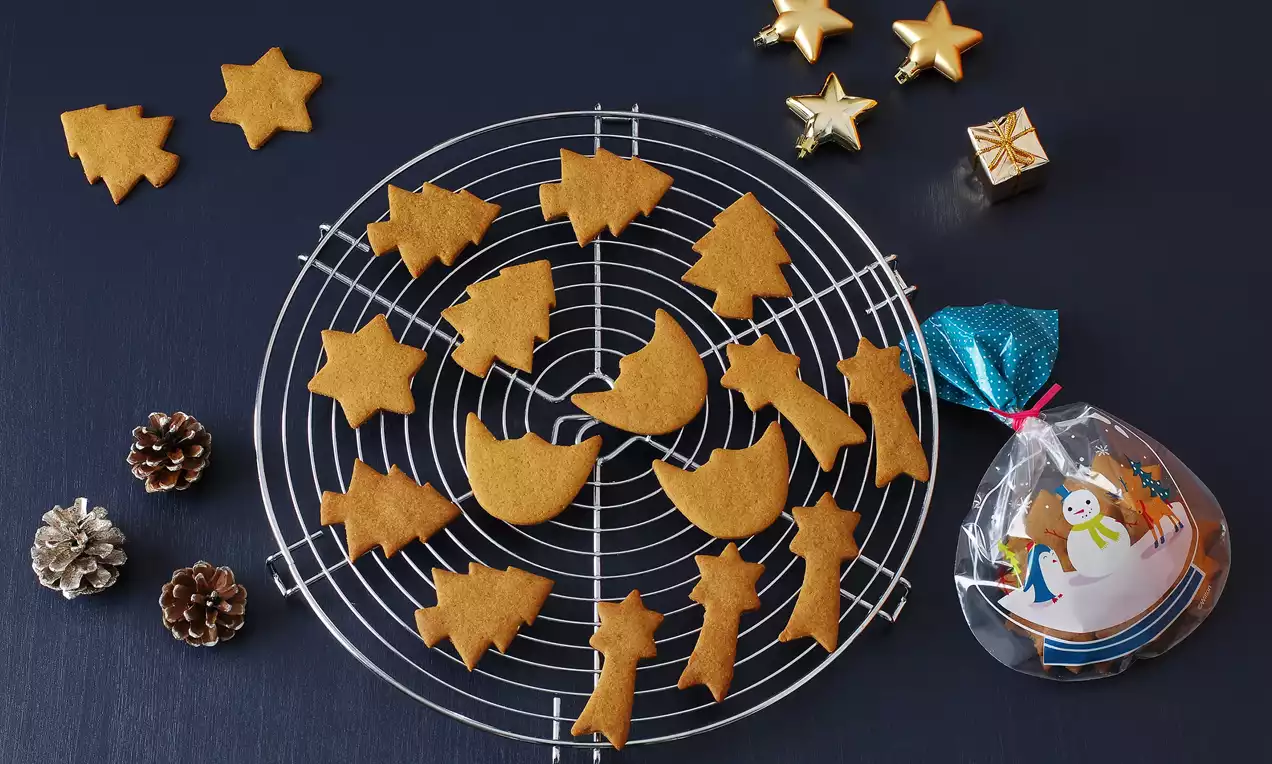 Zencefilli kurabiyeler yıldız, ağaç ve ay gibi şekillerde, gümüş bir ızgara üzerinde, yanında altın yıldız süsleri ve bir hediye paketiyle