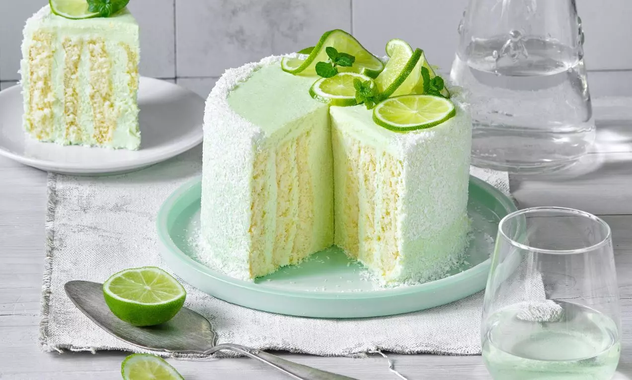 Světle zelený dort s krémovitou texturou, ozdobený limetkovými plátky a mátou, na zeleném talíři s jedním nakrojeným kusem na bílém pozadí