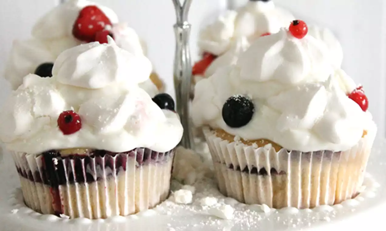 Cupcakes met een goudgele cakebodem, laagjes rood fruit en witte meringue-toefjes, gegarneerd met rode en zwarte bessen op een wit taartrekje
