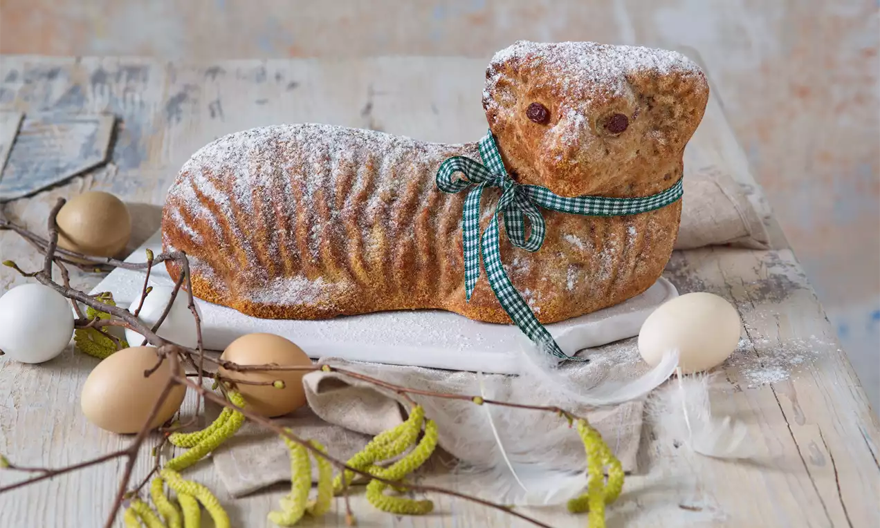 Banánový beránek světle hnědé barvy se záhyby těsta, posypaný moučkovým cukrem, ovázaný zelenou kostičkovanou stuhou, obklopený velikonočními dekoracemi