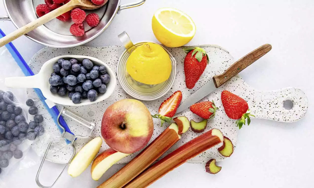 Fruits frais multicolores disposés sur une planche claire avec pommes rouges, fraises éclatantes, rhubarbe rouge, citron et myrtilles