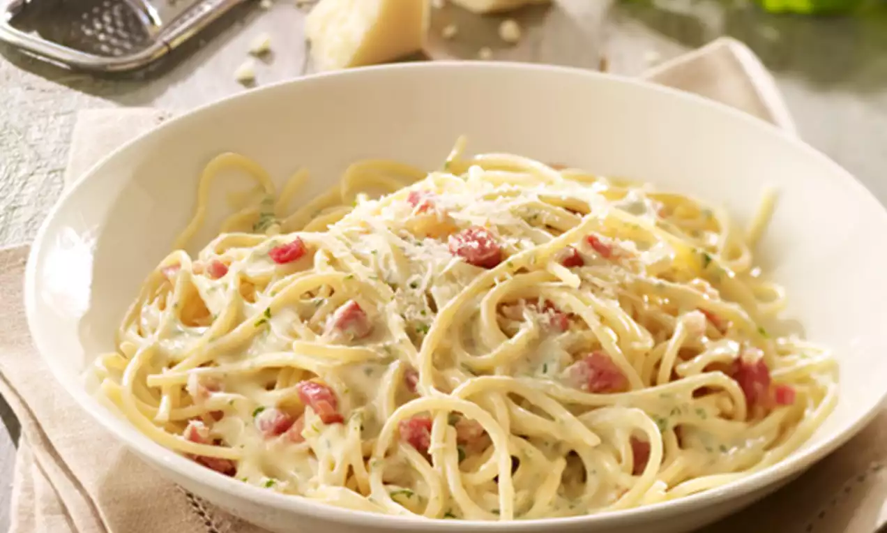Spaghetti in cremiger, hellgelber Sauce mit Speckwürfeln und geriebenem Käse bestreut, angerichtet in einer weißen Schale auf einem beigefarbenen Stoffserviette