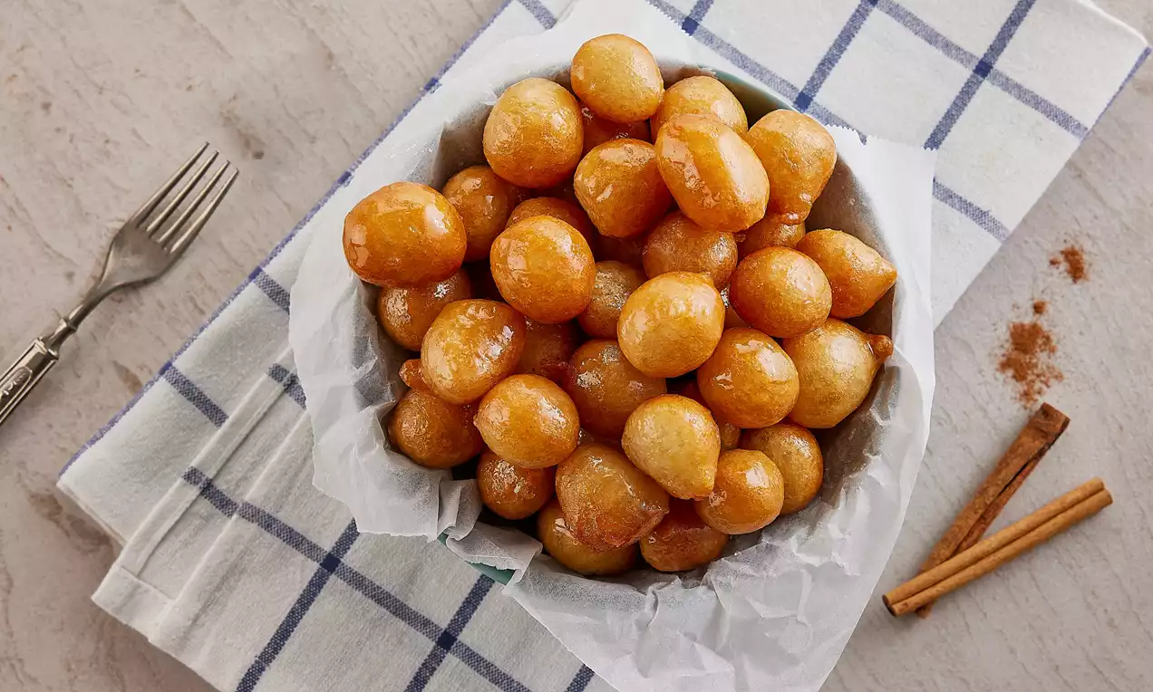 Altın renginde, parlak lokmalar beyaz kağıtla kaplı bir kase içinde, yanında tarçın çubukları ve kare desenli örtü üzerinde sergilenmiş