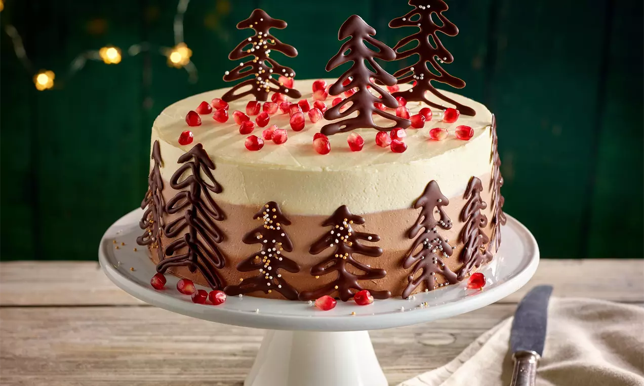 Vánoční dort s vrstvami světle krémové a hnědé náplně, čokoládové stromy, červené kuličky granátového jablka, na bílém podnosu
